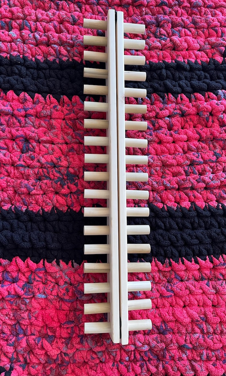 May include: A light-colored wooden drying rack with multiple horizontal dowels. The rack is positioned vertically against a textured background of pink, black, and purple yarn. The rack is made of two vertical pieces connected by the dowels.