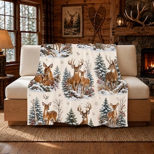 May include: A cream-coloured throw blanket featuring a repeating pattern of deer, including bucks with antlers and fawns, set within snowy winter landscapes with evergreen trees, birch trees, red berries, and small white snowflakes.