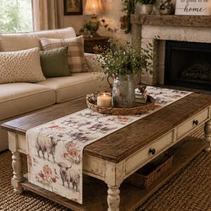 May include: A rustic coffee table with a distressed white base and a dark wood top. A floral and cow-print table runner is displayed on the table, along with a candle and a metal vase. A beige sofa and fireplace are in the background.