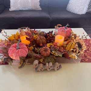 Puede incluir: Un centro de mesa con tem&aacute;tica oto&ntilde;al con calabazas, girasoles, bayas y hojas. Se incluyen dos velas encendidas en portavelas de cristal y un letrero de madera con la palabra "Love".