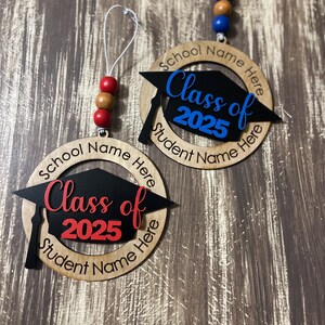 May include: Two wooden graduation ornaments with black graduation caps, red and blue lettering, and the text "Class of 2025". The ornaments have a circle around the cap with the text "School Name Here" and "Student Name Here".