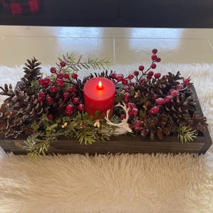 Puede incluir: Centro de mesa rectangular de madera con una vela roja encendida, pi&ntilde;as, bayas rojas y vegetaci&oacute;n. Se incluye una peque&ntilde;a figura de ciervo blanco. El arreglo est&aacute; sobre una superficie blanca y esponjosa.