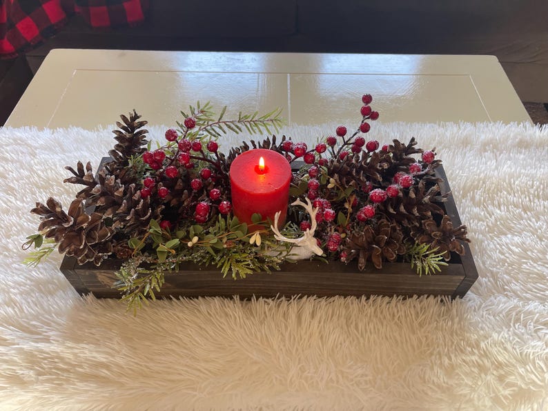 Puede incluir: Centro de mesa rectangular de madera con una vela roja encendida, pi&ntilde;as, bayas rojas y vegetaci&oacute;n. Se incluye una peque&ntilde;a figura de ciervo blanco. El arreglo est&aacute; sobre una superficie blanca y esponjosa.