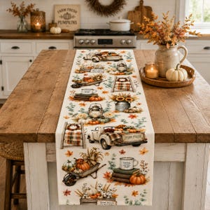 Op de afbeelding: Een crèmekleurige tafelloper met een herfstachtig patroon van vintage vrachtwagens, kruiwagens, lantaarns, stapels boeken, mokken en pompoenen. Het ontwerp bevat oranje, gele en groene accenten van herfstbladeren over de gehele stof.