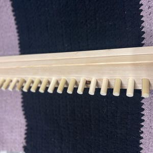 May include: A light-colored wooden shelf with evenly spaced pegs along the bottom edge. The shelf is made of unfinished wood, and the pegs are cylindrical. The background is a black and purple patterned surface.