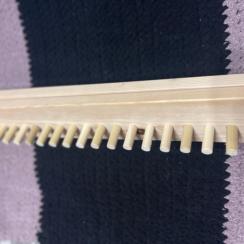 May include: A light-colored wooden shelf with evenly spaced pegs along the bottom edge. The shelf is made of unfinished wood, and the pegs are cylindrical. The background is a black and purple patterned surface.