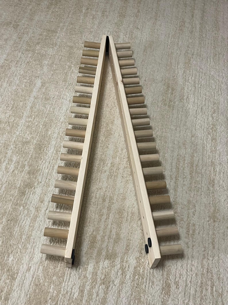 May include: A wooden wine rack with a ladder-like design. The rack is made of light-colored wood and features multiple horizontal dowels for holding wine bottles. The two sides are connected at the top with a hinge, allowing it to stand upright.