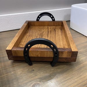 Puede incluir: Una bandeja rectangular de madera con un acabado marr&oacute;n intenso. La bandeja presenta dos asas negras en forma de herradura. La bandeja es de madera y tiene una est&eacute;tica r&uacute;stica y campestre.
