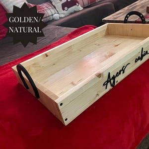 Puede incluir: Una bandeja rectangular de madera clara con asas negras y la inscripci&oacute;n "Ayers cabin" en negro. La bandeja est&aacute; sobre una superficie roja, con el texto "GOLDEN/NATURAL" en la esquina superior izquierda.