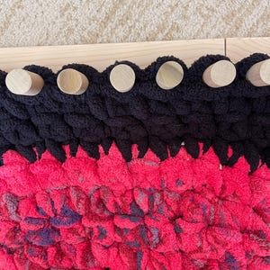 May include: Close-up of a handmade, chunky knit blanket in black and bright pink. The blanket has a scalloped edge with wooden dowels attached to a wooden frame. The pink section has hints of purple.