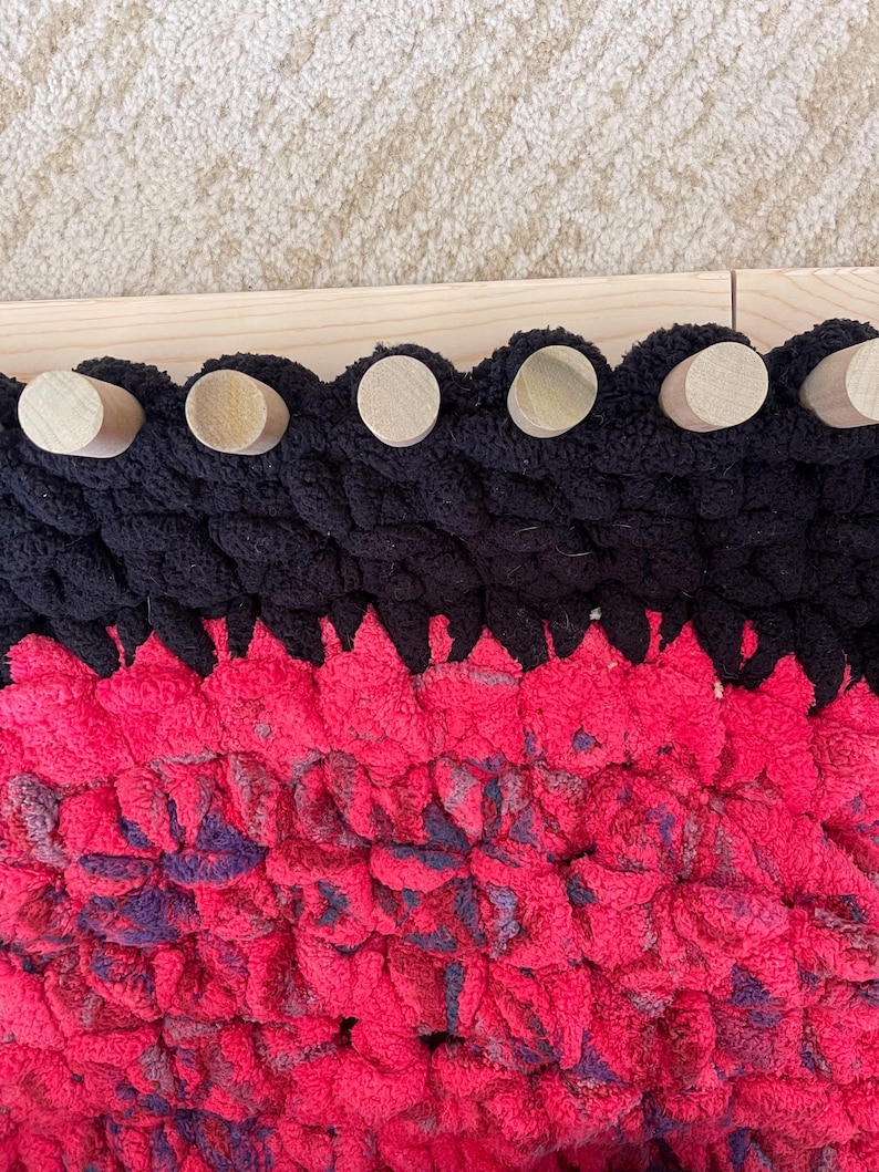 May include: Close-up of a handmade, chunky knit blanket in black and bright pink. The blanket has a scalloped edge with wooden dowels attached to a wooden frame. The pink section has hints of purple.
