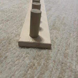 May include: A wooden coat rack with a natural finish. The rack features a rectangular base with evenly spaced, cylindrical pegs. The pegs are made of the same light-colored wood as the base. The rack is designed for hanging coats, hats, or other items.