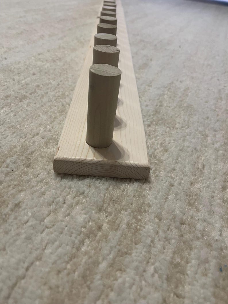 May include: A wooden coat rack with a natural finish. The rack features a rectangular base with evenly spaced, cylindrical pegs. The pegs are made of the same light-colored wood as the base. The rack is designed for hanging coats, hats, or other items.