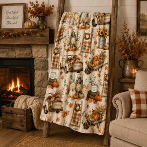 May include: A cream-colored throw blanket featuring a repeating autumn-themed pattern of vintage trucks, wheelbarrows filled with pumpkins, lanterns, sunflowers, ladders, and mugs with the text Farm Fresh, accented by orange and brown fall leaves.