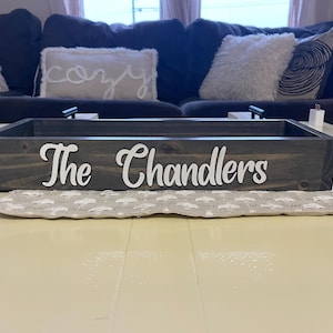 Puede incluir: Una bandeja de madera te&ntilde;ida oscura con las palabras "The Chandlers" en escritura blanca. La bandeja se encuentra sobre una tela gris estampada, con un sof&aacute; oscuro y cojines en el fondo.