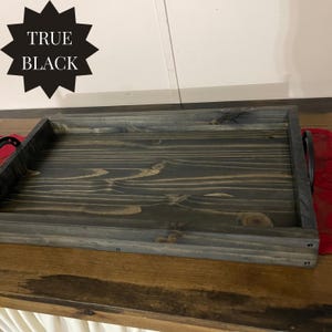 Puede incluir: Una bandeja de madera rectangular con tinte gris oscuro y asas negras. La bandeja tiene un acabado envejecido, mostrando la veta de la madera. El texto "TRUE BLACK" se muestra en forma de estrella negra.