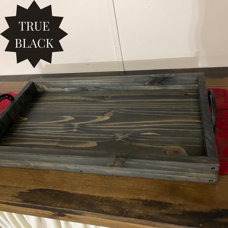 Puede incluir: Una bandeja de madera rectangular con tinte gris oscuro y asas negras. La bandeja tiene un acabado envejecido, mostrando la veta de la madera. El texto "TRUE BLACK" se muestra en forma de estrella negra.