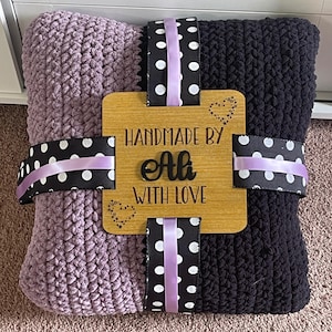 May include: A handmade blanket in shades of purple and black, tied with black ribbon featuring white polka dots and lavender ribbon. A wooden tag reads "HANDMADE BY Ali WITH LOVE" with heart accents.