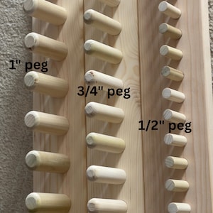 May include: Wooden pegboard with various sizes of wooden pegs. The pegs are labeled as 1", 3/4", and 1/2" in size. The pegs are cylindrical and made of light-colored wood, mounted on a wooden board.