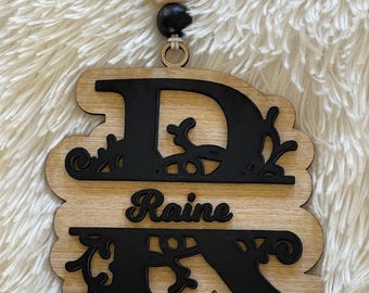 May include: Wooden ornament with the letters "DR" in black with a decorative floral design. The name "Raine" is written in black cursive between the letters.