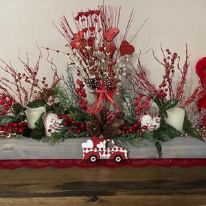 Puede incluir: Un centro de mesa de San Valent&iacute;n en una artesa de madera gris. Presenta bayas rojas, decoraciones en forma de coraz&oacute;n, velas y un cami&oacute;n decorativo. Un oso de peluche rojo es visible a la derecha. El arreglo est&aacute; sobre un camino de mesa rojo.