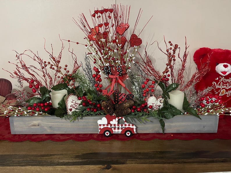 Puede incluir: Un centro de mesa de San Valent&iacute;n en una artesa de madera gris. Presenta bayas rojas, decoraciones en forma de coraz&oacute;n, velas y un cami&oacute;n decorativo. Un oso de peluche rojo es visible a la derecha. El arreglo est&aacute; sobre un camino de mesa rojo.