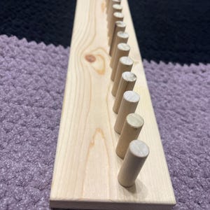 May include: A wooden peg rack with multiple cylindrical pegs. The rack is made of light-colored wood, and the pegs are evenly spaced along its length. The background is a textured purple surface.