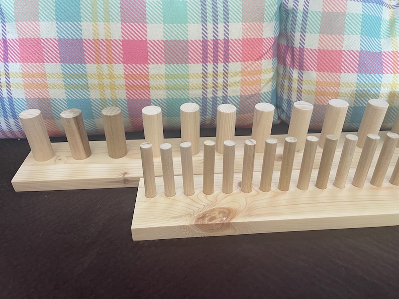 May include: Two unfinished wooden peg racks. One rack has angled pegs, the other has vertical pegs. The racks are made of light-colored wood and are set against a plaid pillow background.