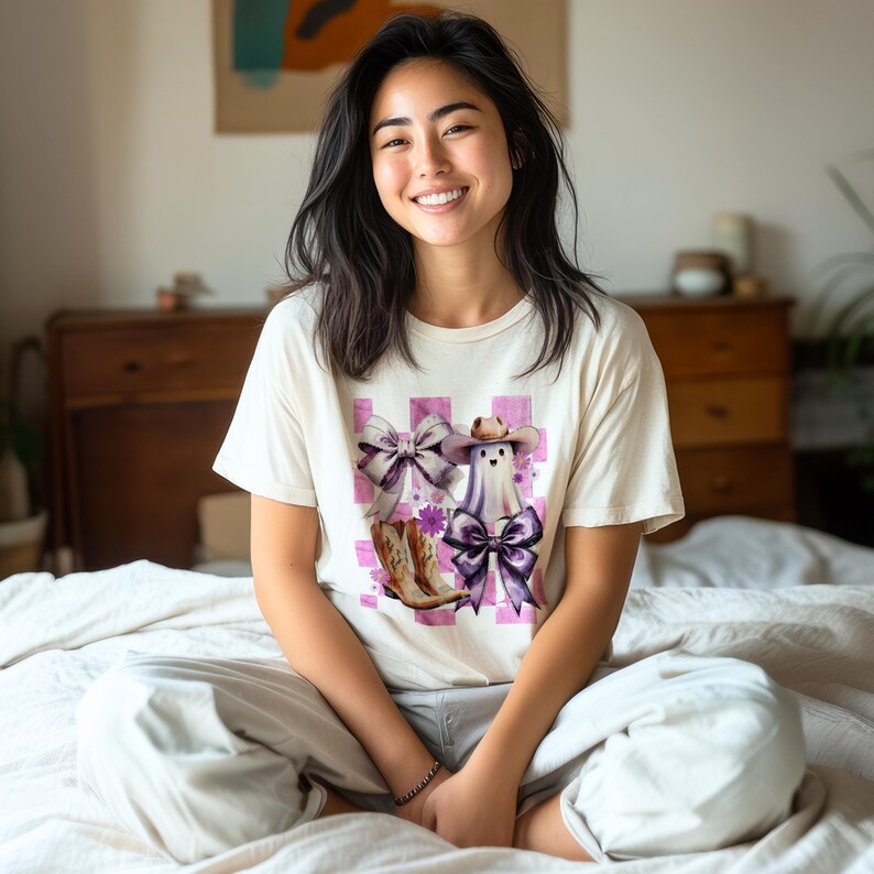May include: Cream-colored t-shirt featuring a whimsical design of a ghost wearing a cowboy hat and a large purple bow, paired with cowboy boots and bows. The design is set against a pink and white checkered background. The shirt is a casual wear item.