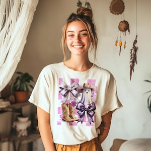 May include: Cream-colored t-shirt featuring a whimsical graphic of a ghost wearing a cowboy hat and boots, with purple bows and a checkered background. The design incorporates floral accents, creating a playful and unique aesthetic.