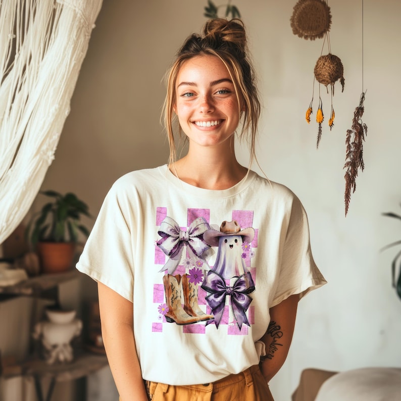 May include: Cream-colored t-shirt featuring a whimsical graphic of a ghost wearing a cowboy hat and boots, with purple bows and a checkered background. The design incorporates floral accents, creating a playful and unique aesthetic.