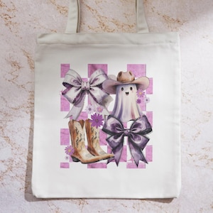 May include: White tote bag featuring a watercolor design of a ghost wearing a cowboy hat and a large bow. The design includes cowboy boots, bows, and a pink checkered background with floral accents. The bag has two beige handles.