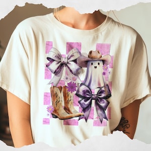 May include: Cream-colored t-shirt featuring a whimsical graphic. The design includes a ghost wearing a cowboy hat, cowboy boots, and purple bows, set against a pink and white checkered background. Daisies add a touch of floral detail.