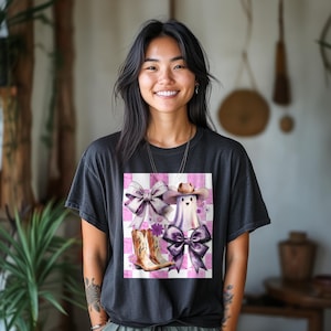 May include: A black t-shirt featuring a graphic design of a ghost wearing a cowboy hat, cowboy boots, and bows on a pink and white checkered background. The design includes purple and white elements.