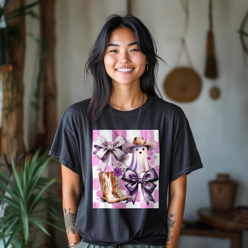May include: A black t-shirt featuring a graphic design of a ghost wearing a cowboy hat, cowboy boots, and bows on a pink and white checkered background. The design includes purple and white elements.
