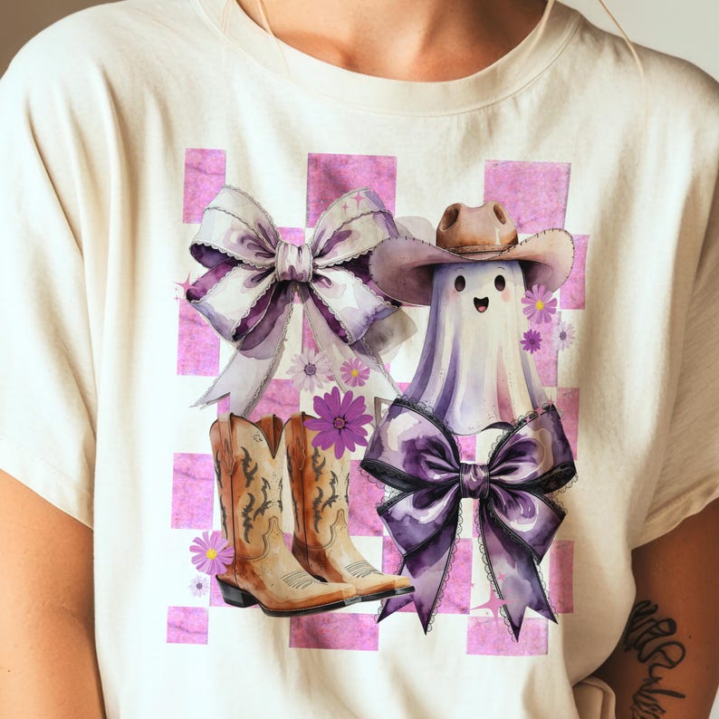 May include: A light-colored t-shirt featuring a watercolor design with a ghost wearing a cowboy hat and a purple bow. The design also includes a pair of cowboy boots, bows, and flowers on a pink checkered background.