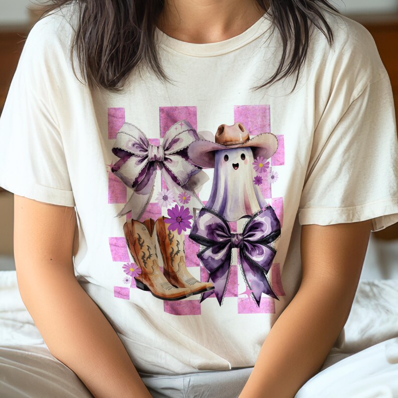 May include: Cream-colored t-shirt featuring a whimsical design of a ghost wearing a cowboy hat and a large purple bow. The design includes a pair of cowboy boots, bows, and flowers, set against a pink and white checkered background.