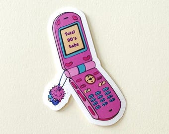 May include: A sticker of a pink flip phone with the text "Total 90's babe" on the screen. The phone has a small pom-pom charm attached. The sticker is on a white background.