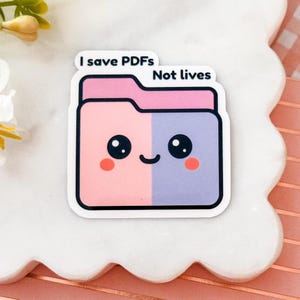 May include: A die-cut sticker shaped like a file folder with a split pink and purple color scheme. The folder has a cute face with large black eyes, a small smile, and pink cheeks. The text "I save PDFs Not lives" is printed in black above.