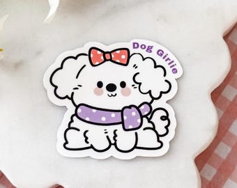 May include: A white cartoon dog sticker with a red polka dot bow on its head and a purple polka dot scarf around its neck. The dog has black outlines, pink cheeks, and the text Dog Girlie printed in purple letters along the upper right side.