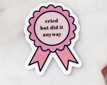 Op de afbeelding: Een roze award lint sticker met een geschulpte rand en twee bungelende uiteinden met kleine oranje stippen. Het midden van het lint bevat de zwarte tekst 'cried but did it anyway' gedrukt in een eenvoudig, vet, schreefloos lettertype.