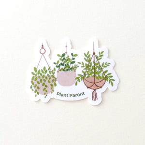 May include: A sticker featuring three hanging plants in pots. The plants have green leaves and are suspended by brown or pink cords. The text "Plant Parent" is printed below the plants.