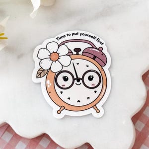 May include: A die-cut sticker featuring a peach-colored alarm clock with a smiling face, round glasses, and a white flower on the side. The clock has a purple top bell and the text "Time to put yourself first" arched along the top edge.