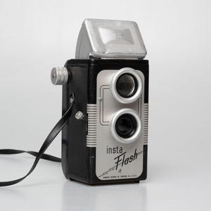 May include: A vintage black and silver Insta Flash camera with a silver flash attachment. The camera has two lenses and a black strap. The text "insta Flash" is printed on the front of the camera.