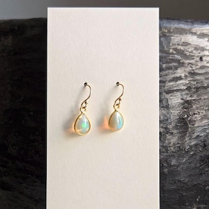 May include: A pair of gold-coloured drop earrings featuring pear-shaped, iridescent opal stones. The earrings are displayed on a white card, with a dark, textured background. The earrings have a simple, elegant design.