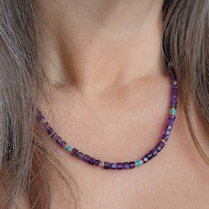 Op de afbeelding: Een ketting met kleine, vierkante, dieppaarse amethiststenen. De ketting heeft kleine turkooizen en goudkleurige kralen ertussen. De ketting wordt om de nek gedragen.