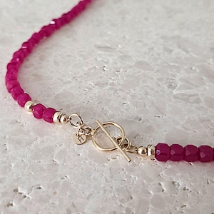 Peut inclure: Un collier composé de petites perles facettées d'un magenta vif. Le bijou est fermé par un fermoir à bascule en métal doré et comporte une petite breloque circulaire gravée de la lettre H près du mécanisme de fermeture.