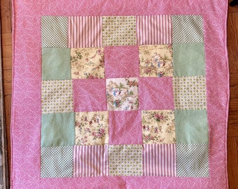 Puede incluir: Una colcha de retazos rosa con una variedad de patrones florales y geométricos en tonos de verde, amarillo y blanco. La colcha está compuesta por nueve cuadrados, cada uno con un diseño diferente.