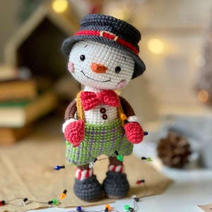 Pode incluir: Um boneco de neve de crochê vestindo um chapéu-coco preto com uma faixa vermelha, uma gravata borboleta vermelha, calças xadrez verdes e roxas e luvas vermelhas. O boneco de neve está segurando uma corda de luzes de Natal coloridas.