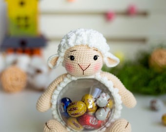 May include: A crocheted lamb-shaped container with a clear center filled with colorful foil-wrapped candies. The lamb has a white head and ears, a beige body, and is sitting on a white surface. Decorative elements are in the background.
